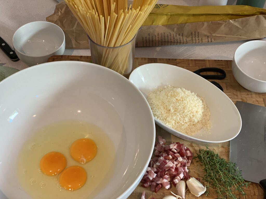 Schüssel mit 3 rohen eiern, gewürfelter Speck, Schüssel mit geriebenem Parmesan, Thymian und Spaghetti